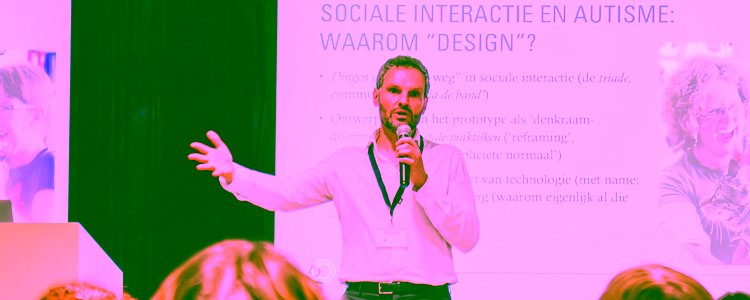 Project Jelle van Dijk gives a presentation on stage about the relevance and importance of design research in the context of autism. He is standing in front of an audience, holding a microphone.
