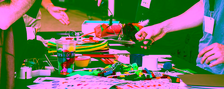 Four people are standing around a table with creative colourful tinkering materials, such as building blocks, fluffy straws, and stickers.